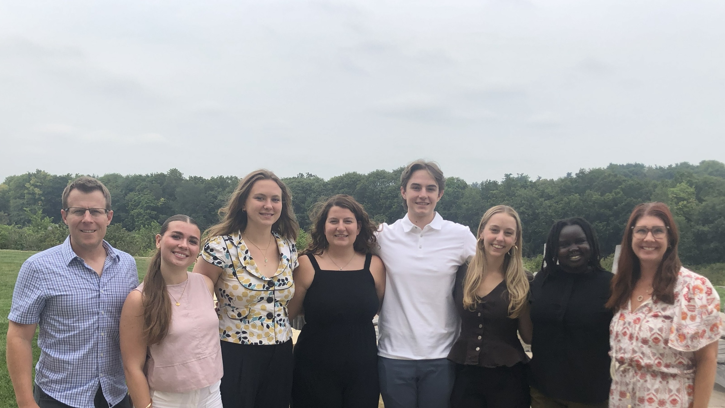 A group of eight people smiling outdoors with trees and a cloudy sky in the background.